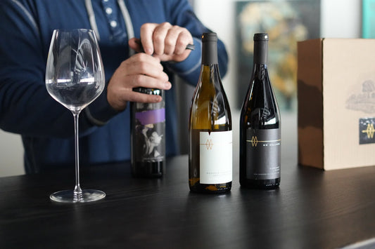 An empty wine glass next to three wine bottles on a table. One bottle is held being opened. 